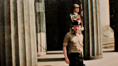 STEPHEN CLOSE An archive picture of a soldier posing at the camera. He is a beige military uniform shirt with a beret on his head. He is stood in front of a building fronted by large pillars. In the background you can see a guardsman holding a rifle looking away from the camera.