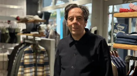 Shop owner Rick Gaglio, who has dark hair and is wearing a black shirt, stands in front of men's clothing on shelves and on hangers