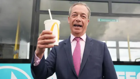 PA Media Nigel Farage smiling with his mouth open while holding a McDonald's milkshake. He is wearing a navy suit jacket and pink shirt, with a pink tie.