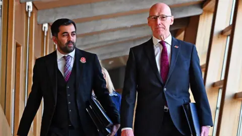 Getty Images Humza Yousaf, with dark hair and wearing a dark suit and purple tartan tie, walks in the Scottish Parliament alongside John Swinney, a bald man with glasses wearing a dark suit and purple tie. 