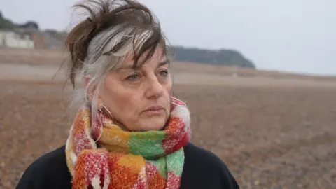 Rebecca Sinker, wearing a red, green and orange striped scarf, standing on a beach