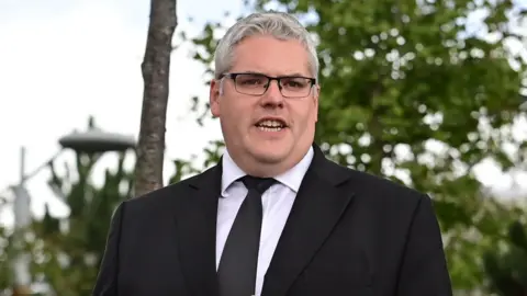 Pacemaker press Gavin Robinson has short grey hair and black framed glasses. He is wearing a black suit, dark plain tie and white shirt. Trees are in the background.