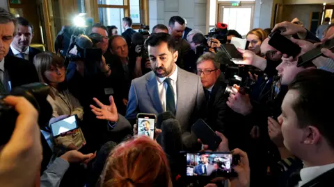 PA Media Humza Yousaf standing in the middle of a large scrum of journalists at Holyrood