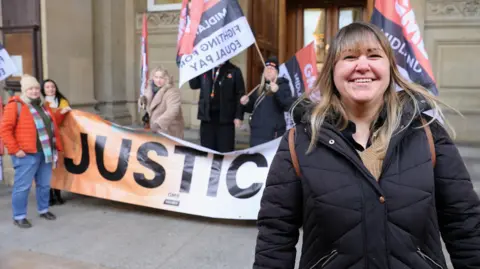 GMB Union A woman in a black puffa coat standing outside a sandstone building with ornate carving on some bricks and stone pillars. Behind her stand four people holding a banner and flags saying 