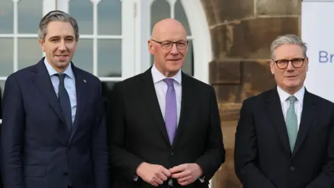 ROBERT PERRY/POOL/EPA-EFE/REX/Shutterstock Irish Taoiseach (PM) Simon Harris, First Minister of Scotland John Swinney and British Prime Minister Sir Keir Starmer pose for a photo at the British-Irish Council summit in Edinburgh.  All three men are wearing dark-coloured suits. 