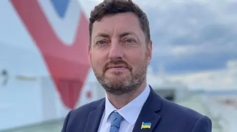 BBC Cammy Day, with dark hair and wearing a dark blue suit with a Ukrainian badge attached, looks at the camera in a close-up shot with a blurred background 