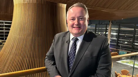 BBC Darren Millar is stood slighting to the right of the middle of the shot, on a balcony overlooking the funnel in the middle of the Senedd building. He is in a suit, tie and white shirt, and is smiling.