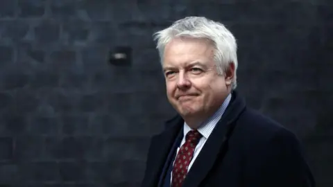 Getty Images Carwyn Jones, who has grey hair, wears red patterned tie, vertically striped shirt and a dark coat. Behind him is a grey brick wall