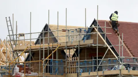 PA Media A construction site showing a house in progress, with scaffolding surrounding the structure and a worker in a high-visibility vest installing roof tiles on the sloped roof. Wooden beams and other construction materials are visible in the foreground and background