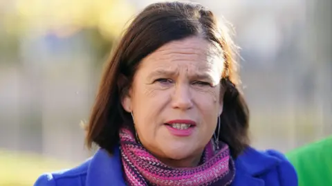 PA Media Mary Lou McDonald looking straight at the camera. She is wearing a blue coat and pink scarf. She had dark brown hair and pink lipstick.