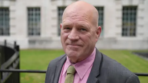 ANN GANNON/BBC Stephen Close smiles at the camera in front of a green grassy lawn, black railings and a light-coloured-brick building in Whitehall, central London. He wears a pink shirt, sage green tie and a grey suit.