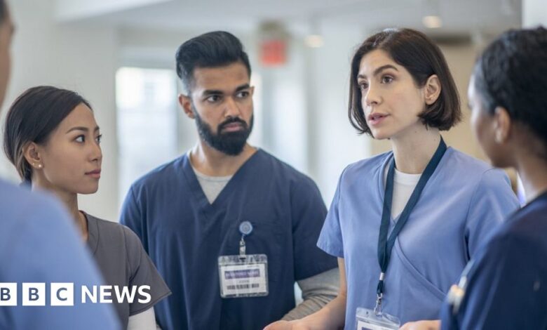 A group of five healthcare workers in conversation in a hospital. The three women and two men are wearing blue scrubs and ID badges.