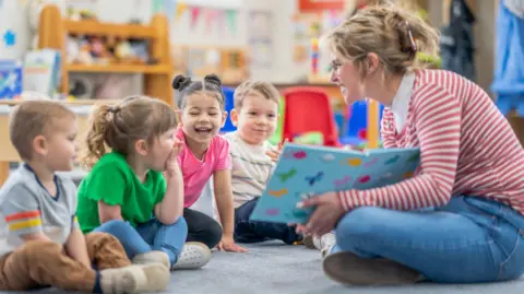 Getty Images A woman reads a book to four young children in a nursery room. She is sat cross-legged on the floor with a blue book open to the two girls and two boys, who are smiling.