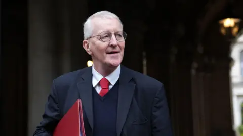 PA Media Hilary Benn stands in holding a red folder. He has glasses and grey hair. He is wearing a black suit with a red tie.