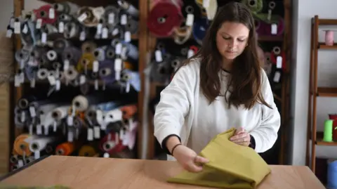 Getty Images A woman wearing a white jumper working at a fabric shop in east London. She is folding some yellow material on a wooden counter, with rolls of material seen stacked behind her