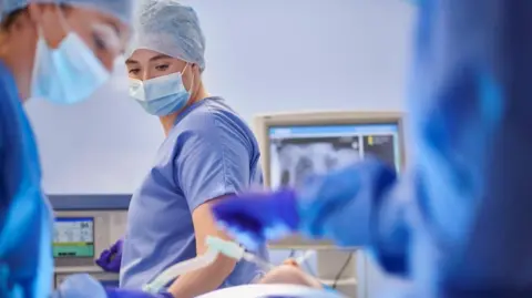 Getty Images A woman operating a machine in an operating theatre, wearing blue uniform and a surgical mask. She is in front of a person being operated on with a tube in their mouth.