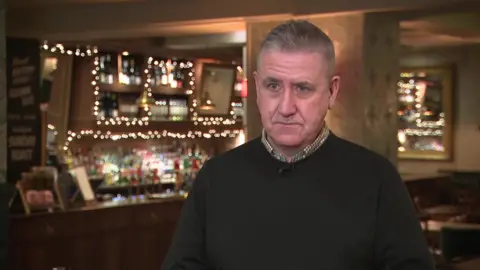 Mick Dore who has grey hair and blue eyes, standing with the bar of the Alexander pub in Wimbledon behind him, wearing a black jumper with a brown and white checked shirt collar