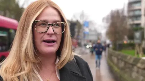 Kaylan Evans with long blonde hair, a pair of glasses, standing on a street with a bus in the background.