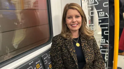 LDRS Kim McGuinness is sitting next to a window on a train on the Tyne and Wear Metro. She has blonde hair and is wearing a large gold pendant and is wearing a leopard skin jacket