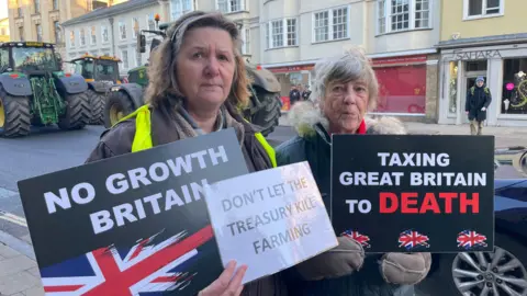 BBC/Malcolm Prior Caroline Graham and fellow protestor holding signs saying No Growth Britain, and Don't let the Treasury kill farming