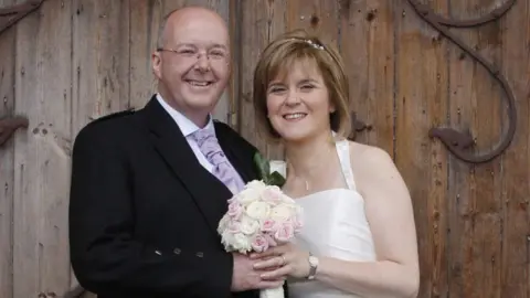 PA Media Nicola Sturgeon and Peter Murrell pictured on their wedding day in front of a wooden door. Sturgeon is wearing a white wedding dress, with Murrell wearing a dark jacket and purple tie. They are holding a bouquet of flowers.