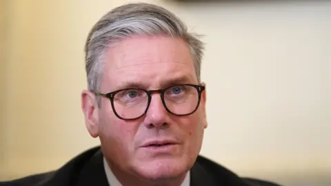 PA Media Sir Keir Starmer is pictured sitting in a room looking away from the camera. He is wearing black glasses and a black suit with a white shirt underneath