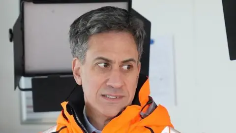 John Fairhall/BBC Ed Miliband looks away from the camera. He has dark short hair with some grey in it. He is wearing a bright orange coat with a shirt just visible underneath