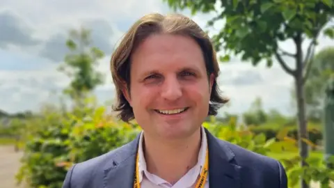 Paul Moseley/BBC Liberal Democrat MP Steff Aquarone, a man with shoulder length, wearing a blue suit jacket and open necked shirt smiles for the camera on a sunny day