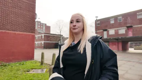 A blonde woman wearing a jacket walks through a housing estate