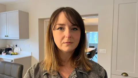 Crystal pictured indoors in her kitchen; she's wearing a grey-black denim jacket and is looking directly into the camera with a sombre expression. 