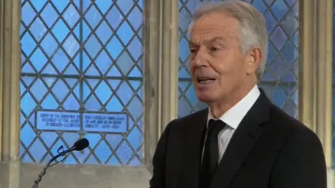 Former PM Tony Blair with grey hair mid-speech as he deliveries a eulogy for John Prescott. There is a microphone in front of him. He is wearing a black suit and tie