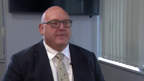 Picture of Vincent Hodder in a blue suit jacket and white shirt.
He is wearing a brown tie with blue and white dots on and glasses. 