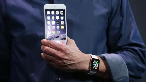 Getty Images Close up image of a man's hand holding a white iPhone 6, with an Apple Watch on his wrist. His dark blue shirt is rolled up at the cuff.