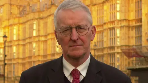 BBC A man look at the camera, he has short grey hair and round glasses. He is wearing a suit with a red tie and white shirt and dark overcoat. He is standing outside in front of the Houses of Parliament in London. 