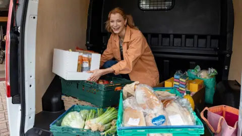 Getty Images Volunteer looking at the camera laughing while unloading a van filled with food in trays for a food bank. She is in the North East of England and is happy to be giving back to her community.