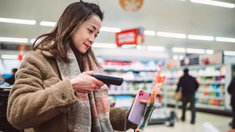 Getty Images Young woman with dark hair, wearing brown fake fur jacket and grey and pink scarf scans packet of herbs at supermarket  