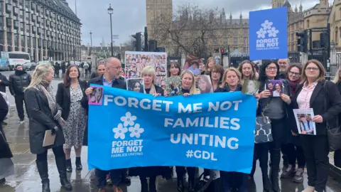 BBC A gathering of men and women with a large blue flag in front of the Houses of Parliament. The flag reads forget me not families uniting.