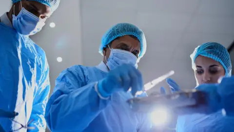 Getty Images Three surgeons wearing face masks, gloves and blue scrubs look down at some surgical tools in preparation for an operation.