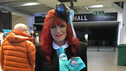 BBC Gloria Jane Martin wears a black suit, a turquoise tie with a Reform UK badge pinned on and a black fascinator with another badge and Reform rosette attached,