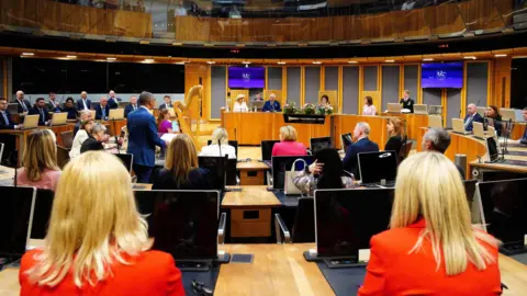 Reuters A general view of the Senedd during a royal visit, with the King and Queen Camilla sat in the centre, and a harp in view. Vaughan Gething, who was first minister at the time, is stood with  his back to the camera.