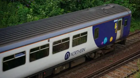 PA A Northern Trains carriage with its white and blue colours on a railway track with thick trees behind.