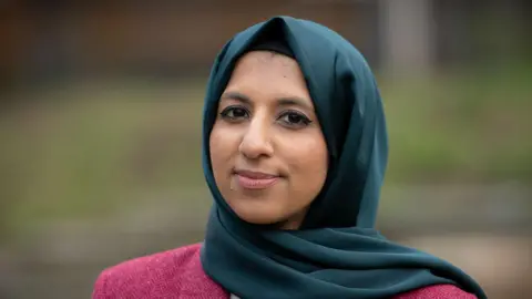 BBC/Emma Lynch Zara Mohammed looks into the camera with a small smile. She wears a dark pink jacket and a teal green hijab.