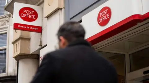 Getty Images Man wearing a dark coat walks by Post Office signage in London