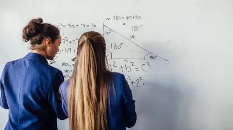 Getty Images Two pupils wearing navy blue blazers face away from the camera as they write algebra on a whiteboard. The student on the left has her brown curly hair in a bun, and her classmate on the right has long light brown hair, half tied up.