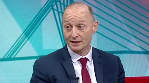 BBC Paul Waugh on the set of Politics North West. He is wearing a dark suit jacket, white shirt and red tie and has short, grey receding hair.