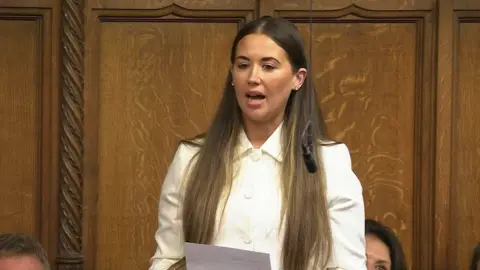 PA Media Sorcha Eastwood speaking in the house of commons. She is wearing a white top and holding a piece of paper in front of her.