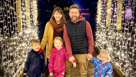 Katie and Tom The family stands under an archway of fairy lights. Katie and Tom are with their three children, a boy and two girls.