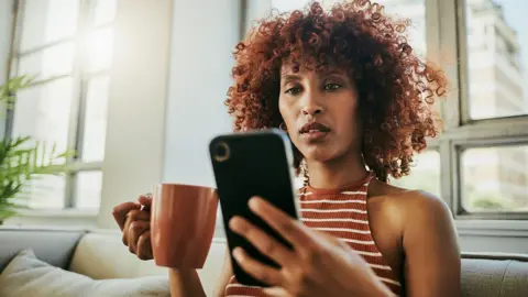 Getty Images A woman looks at her phone with a mug in hand inside her home