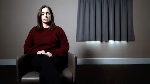 BBC Business coach Rebecca Amin sits on a leather chair. She is wearing a maroon sweater with buttons on the shoulder. Behind her there is a window with curtains pulled shut.
