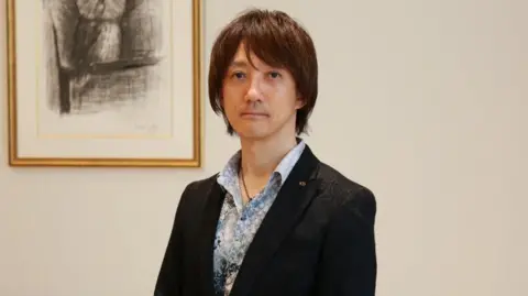 Koei Tecmo Tomohiko Sho stands in front of a wall with a picture of an owl. Mr Sho is wearing a blazer over a colourful blue shirt with an open top button. He is Japanese. He has a neutral expression and smart medium-length hair.